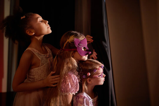 Side view portrait of three little girls wearing costumes peeking over curtain backstage in theater copy space