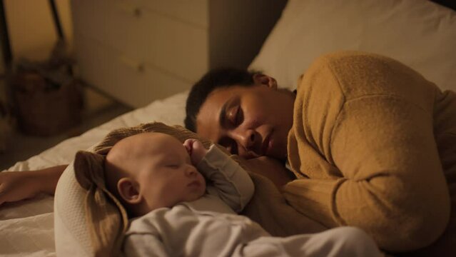 Medium shot of newborn baby boy in bodysuit sleeping in cocoon pod, and tired mother in woolly cardigan lying on bed beside him and taking nap, in dimly lit bedroom at nighttime