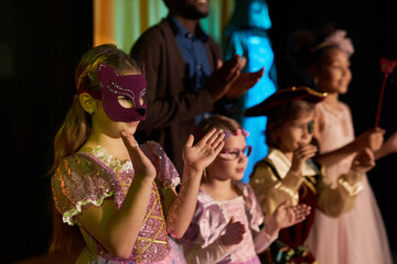 Portrait of teen girl wearing mask and costume standing on stage in theater and applauding with...