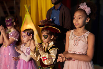 Portrait of Black teenage girl wearing pink princess dress standing on stage in theater with group...