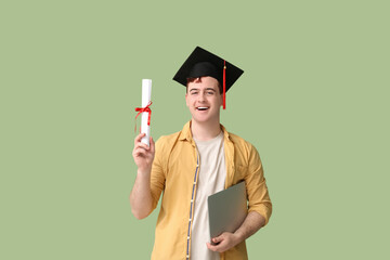 Male student in mortar board with diploma and modern laptop on green background