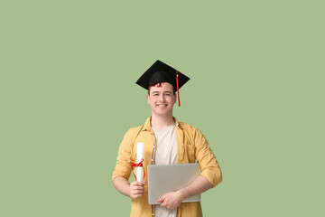 Male student in mortar board with diploma and modern laptop on green background