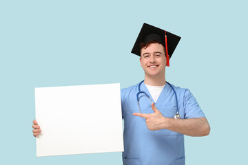 Male student in mortar board and uniform of doctor pointing at blank poster on blue background