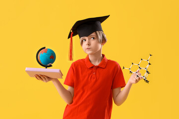 Thoughtful little boy in graduation hat with globe and molecular model on yellow background. End of...