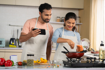 Fototapeta na wymiar Joyful Indian couples cook or trying new food recipe by watching online videos from Mobile phone at Kitchen - concept of technology, relationship bonding and collaboration