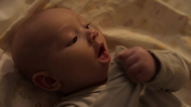 Close-up top shot of face of 3-month-old baby boy lying on back on bed sheet, looking around, flailing arms and chewing on bodysuit sleeve during teething