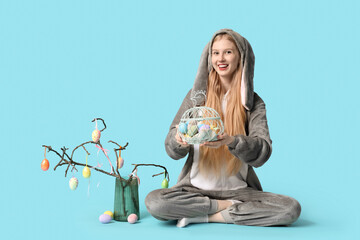 Beautiful young happy woman in bunny costume and tree branches with eggs sitting on blue background. Easter celebration