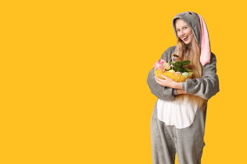 Beautiful young happy woman in bunny costume with basket full of eggs and flowers on yellow background. Easter celebration
