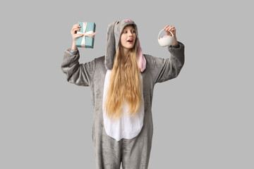 Beautiful young shocked woman in bunny costume holding gift box and basket with egg on grey...