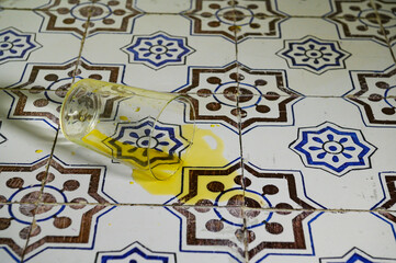 "A clear glass fallen with orange juice on the kitchen floor, tiled with white ceramic tiles adorned with blue, indigo, and brown geometric patterns. These are antique ceramics from the early 20th 