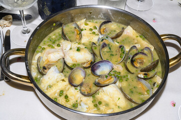"Top view of an aluminum casserole containing freshly made cod in green sauce with clams and peas, ready to be served and enjoyed. White cod pieces, open clams, all surrounded by a creamy sauce