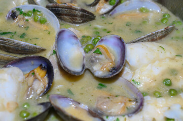 "Close-up of the contents of a casserole with cod in green sauce with clams. In the foreground, an opened clam sits atop the rest, surrounded by other clams, pieces of cod, and scattered peas 