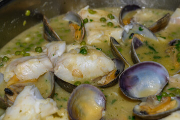 "Close-up of the contents of a casserole with cod in green sauce with clams. In the foreground, an opened clam sits atop the rest, surrounded by other clams, pieces of cod, and scattered peas in the s