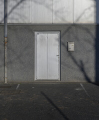 "Close-up of the closed metal entrance door with a gray facade, an empty parking lot, and a mailbox. The facade casts shadows from the branches of a tree overhead.