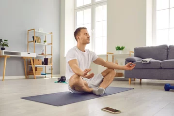 Schilderijen op glas Attractive man in sports outfit doing yoga and meditating on exercise mat. Sporty peaceful calm young man with closed eyes practicing yoga in lotus pose at home. Healthy lifestyle concept © Studio Romantic