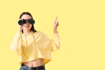 Young woman with binoculars pointing at something on yellow background