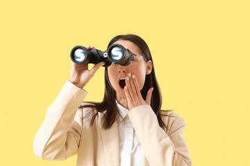 Shocked young businesswoman looking through binoculars on yellow background, closeup