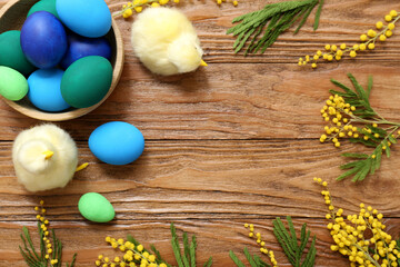Frame made of chicks and festive basket with painted Easter eggs and mimosa flowers on wooden background