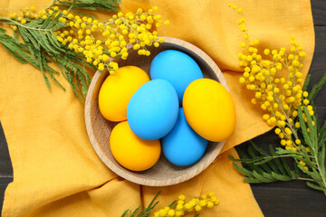 Obraz na płótnie Canvas Basket with painted Easter eggs and mimosa flowers on napkin on dark wooden background