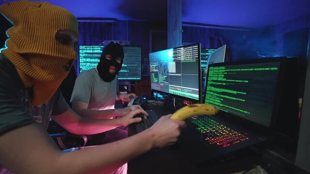 Hacker coding or listening music on programming computer at desk. Male danger individual computing on notebook or hacking pc in neon color light. IT cybercriminal in thief mask hacks encryption access