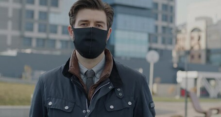 Slow motion portrait of young man putting on face mask and looking at camera standing outdoors in city street. Pandemic and smog pollution concept.