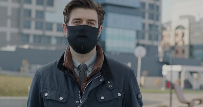 Slow motion portrait of young man putting on face mask and looking at camera standing outdoors in city street. Pandemic and smog pollution concept. - Powered by Adobe