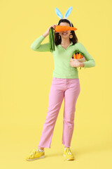 Beautiful young woman in bunny ears with toys carrots on yellow background. Easter celebration