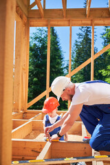 Father with toddler son building wooden frame house. Male builder playing with kid on construction site, wearing helmets and blue overalls on sunny day. Carpentry and family concept.