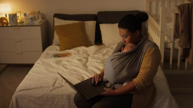 Medium shot of young multiethnic mother with baby son or daughter in sling wrap sitting on bed at home, typing on laptop and writing report, while working part-time during maternity leave