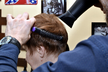 A woman sits in a chair, and a hairdresser uses a comb and a hair dryer to add volume to her hair,...