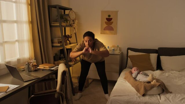 Full shot of young body positive Hispanic or African American mother exercising alone at home and following fitness video on laptop, while baby son is lying in cocoon pod on bed