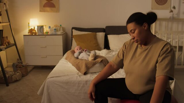 Medium shot of Hispanic or African American woman sitting on fitness ball in bedroom, doing neck rotation and stretch exercise, bouncing then looking at baby lying on bed in cocoon and smiling