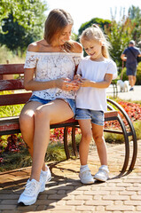 Mother and daughter sitting on a bench in the sunny summer city park. Childhood, leisure and people concept - happy family rest and have a good time