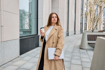 Obraz premium Amidst the city's vibrant energy, a businesswoman, wrapped in a trendy trench coat, gracefully walks along the streets, multitasking with her laptop