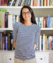 Woman, portrait and happy by bookshelf in house with books for reading, learning and knowledge in living room. Person, face and confidence by home library with magazine, journal and study collection