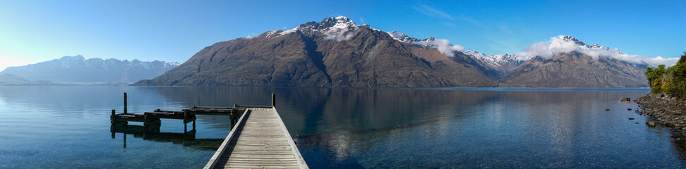 Panoramic view of wooden pier jutting into placid and crystal clear Lake Wakatipu towards Cecil...