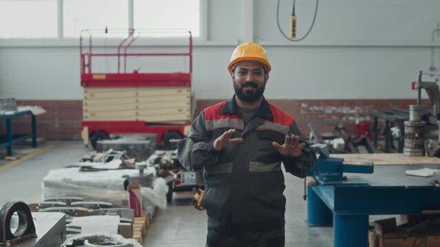 Medium shot of Middle Eastern male engineer technician wearing coverall uniform and yellow hard hat walking along production plant and speaking at camera while giving orientation tour