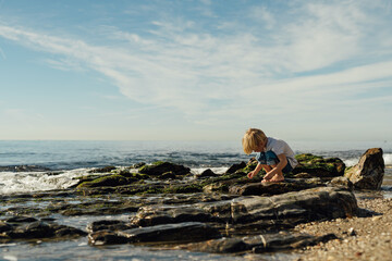 little blond boy playing on the seashore. child playing on the seashore. boy stands on stones in the water. child playing on the seashore