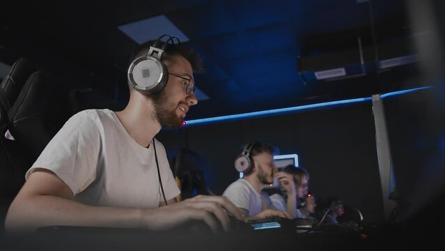Bristly smiling adult gamer in headset playing PC video game at computer club with team cyber sport tournament. E-sport championship. Five player in headphones. Multiplayer online strategy shooter