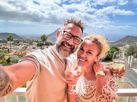 One happy adult couple taking selfie picture at home in terrace with city and mountains outdoors view. People enjoying vacation and leisure together smiling and drinking wine. Joyful people holiday