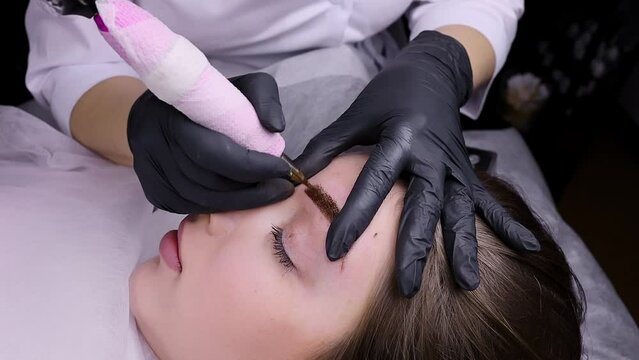 A permanent makeup artist applies strokes to the eyebrow, performing permanent makeup. Permanent makeup procedure, performing PMU of eyebrows