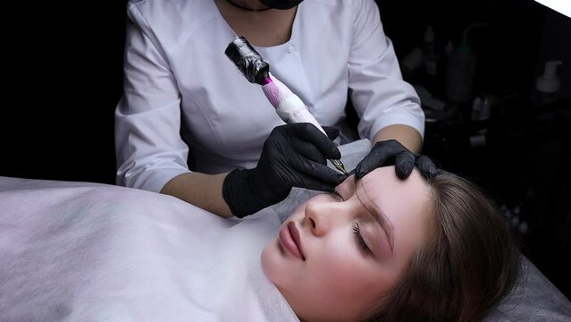 On a dark background, a master in white uniform performs permanent eyebrow makeup procedure. Permanent makeup procedure, performing PMU of eyebrows