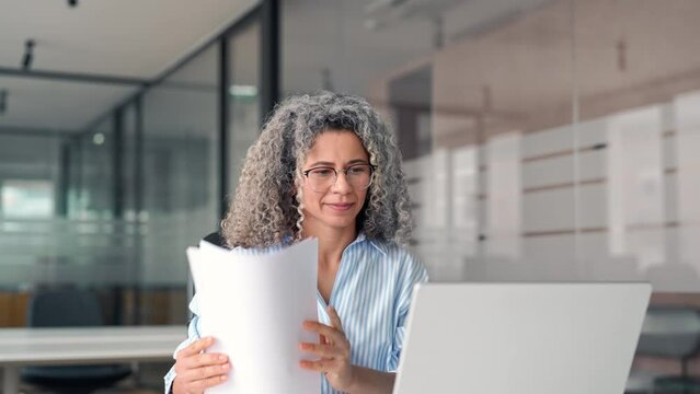 Busy mature business woman working in office looking at laptop holding document. Older professional hr manager, lawyer or notary checking financial tax report account in office. Business consultancy.