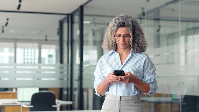 Busy middle aged business woman holding mobile cell phone using cellphone in office. Mature old professional lady business owner entrepreneur using smartphone standing at work.
