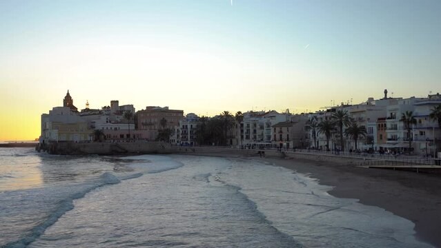 Beautiful city of Sitges at sunset. Orange sky. Camera panning right. San Sebastian beach, in background is the Cathedral of Sitges and old town center.  Travel destination in Catalonia, Spain.
