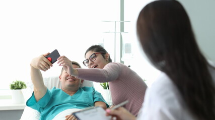 Husband and wife take selfie in hospital bed. Doctor motivates patient. Single room is equipped...