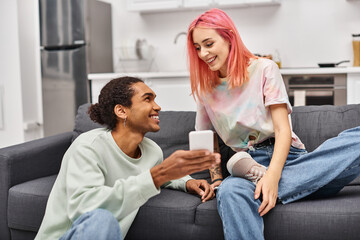 Fototapeta premium beautiful joyful multiracial couple in homewear sitting in living room and looking at phone happily