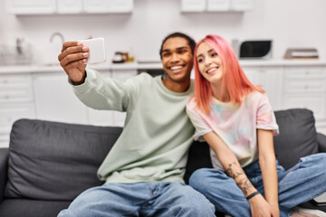 focus on smartphone in hands on jolly diverse couple taking selfies while in living room at home