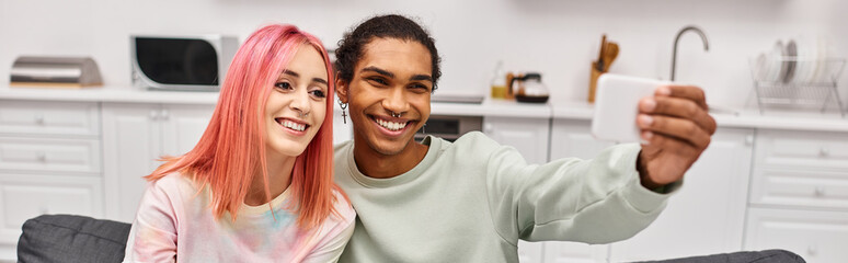 cheerful attractive multicultural couple taking selfies while in living room at home, banner
