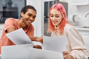 attractive joyous multiracial couple in cozy homewear looking at paperwork while working remotely